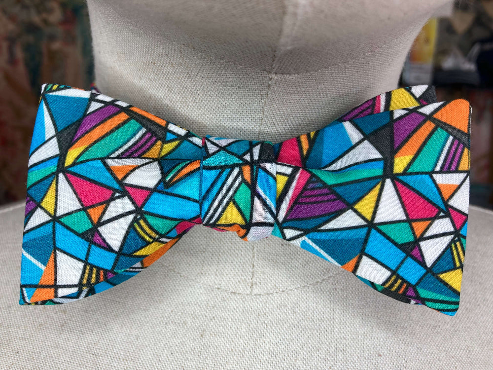 A multicolor geometric patterned bow tie displayed on a light gray fabric mannequin torso. The pattern features a vibrant mosaic of triangles and polygons in blue, pink, orange, yellow, green, purple, and white with black outlines.