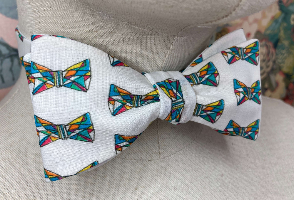 A white self-tie bow tie with a colorful geometric bow tie pattern, displayed on a beige fabric mannequin torso.