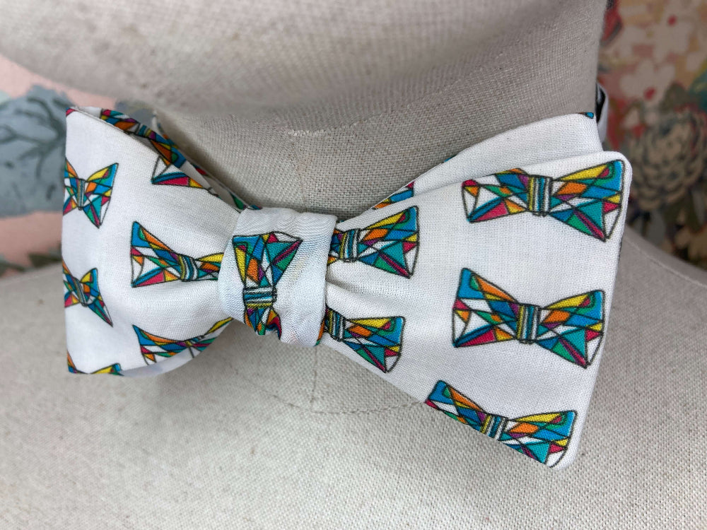 A white self-tie bow tie with a colorful geometric bow tie pattern, displayed on a beige fabric dress form.