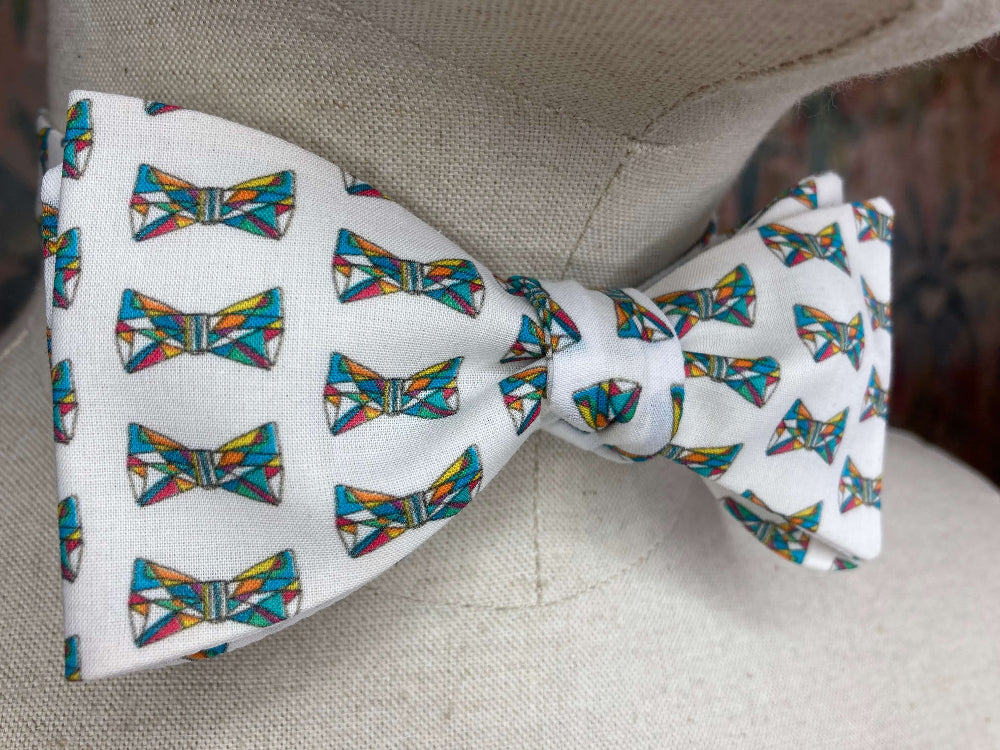 A white self-tie bow tie with a repeating pattern of colorful geometric bow ties, displayed on a beige fabric mannequin torso.