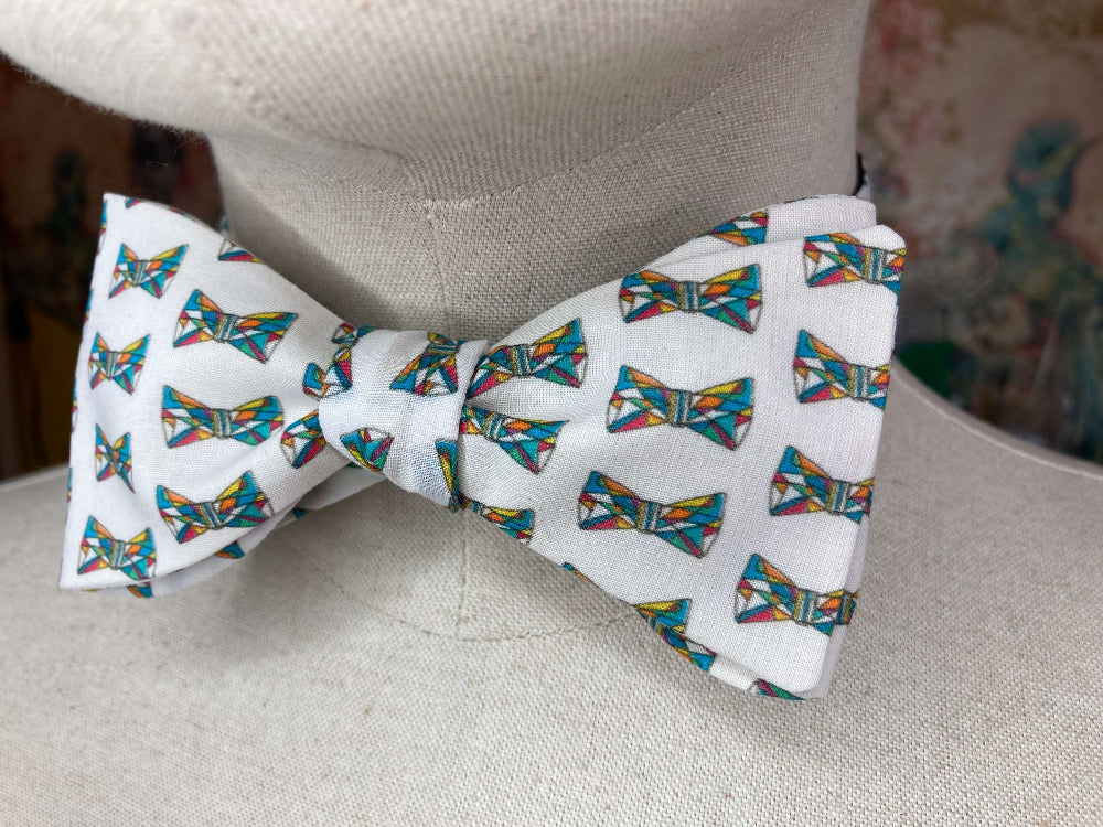A white self-tie bow tie with a colorful geometric bow tie pattern, displayed on a beige fabric mannequin torso.