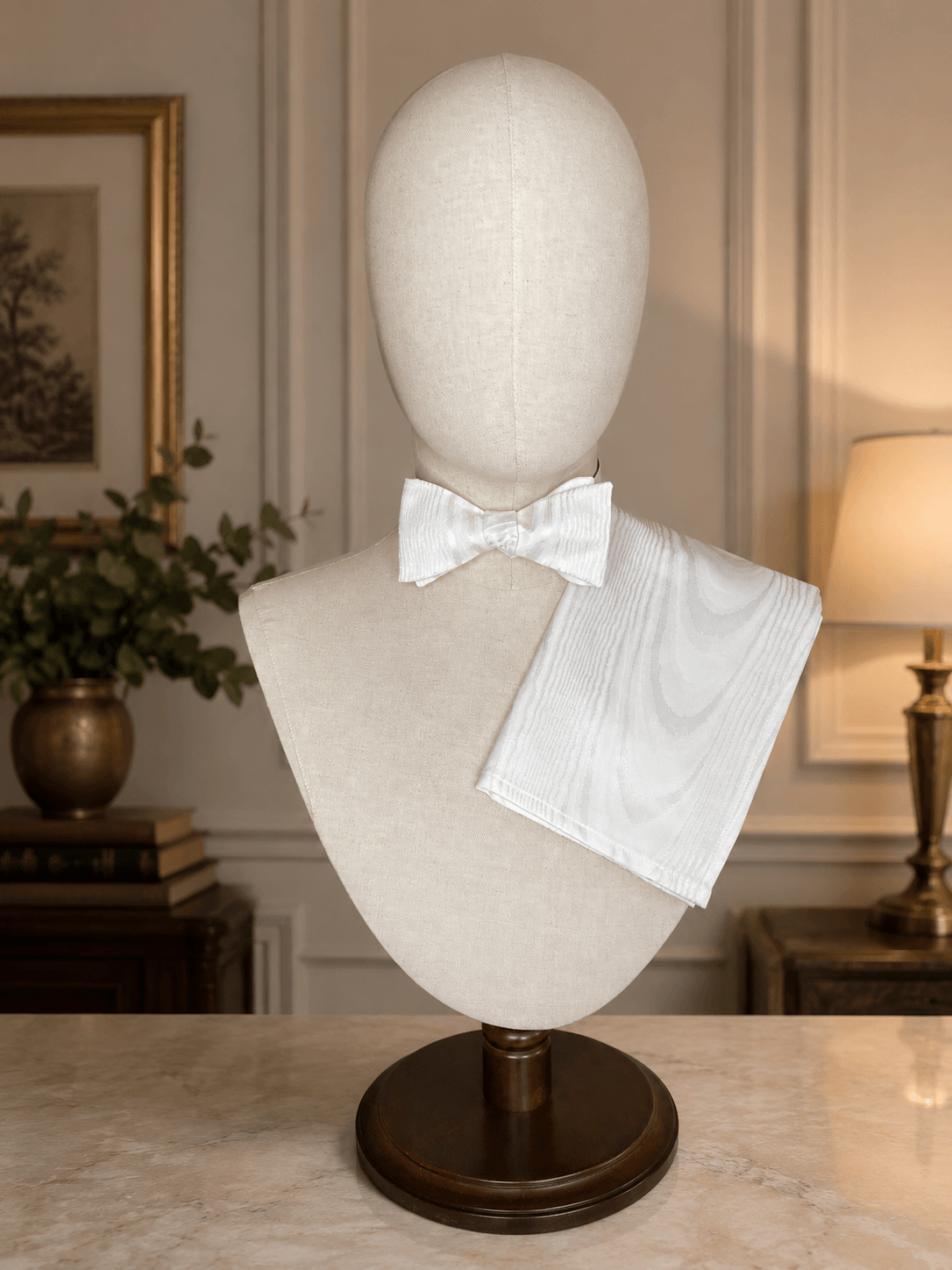 Mannequin head with white bow tie and pocket square on a wooden stand in a room setting.