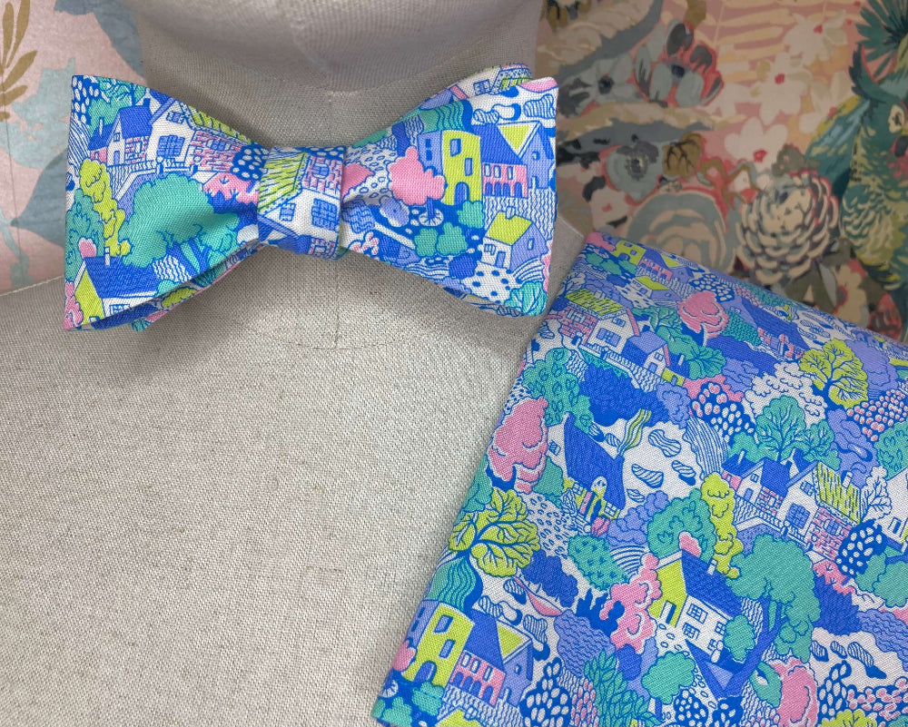 A multicolor patterned bow tie displayed on a beige fabric mannequin, with a matching pocket square laid next to it on a neutral surface.