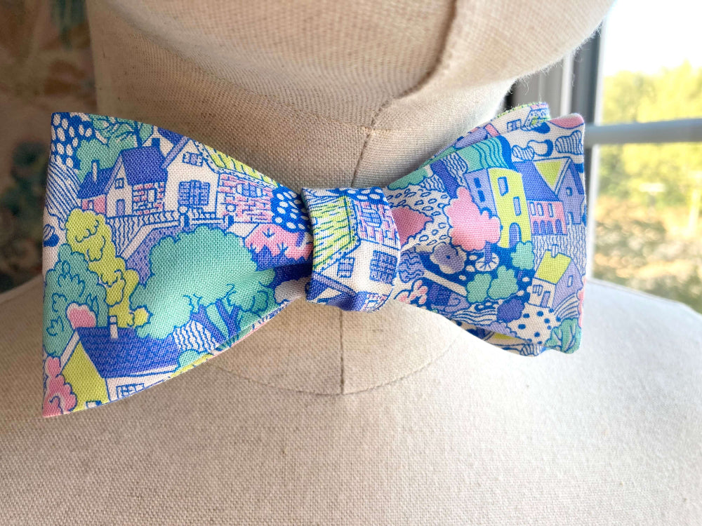 A men's self-tie bow tie with a colorful, whimsical landscape print featuring houses, trees, and clouds in shades of blue, pink, green, and yellow, displayed on a beige fabric mannequin torso.