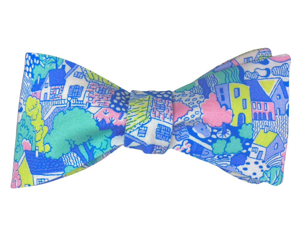 A self-tie bow tie featuring a vibrant, multicolor cartoon landscape print with houses, trees, and clouds in shades of blue, pink, green, yellow, and white, set against a plain white background.