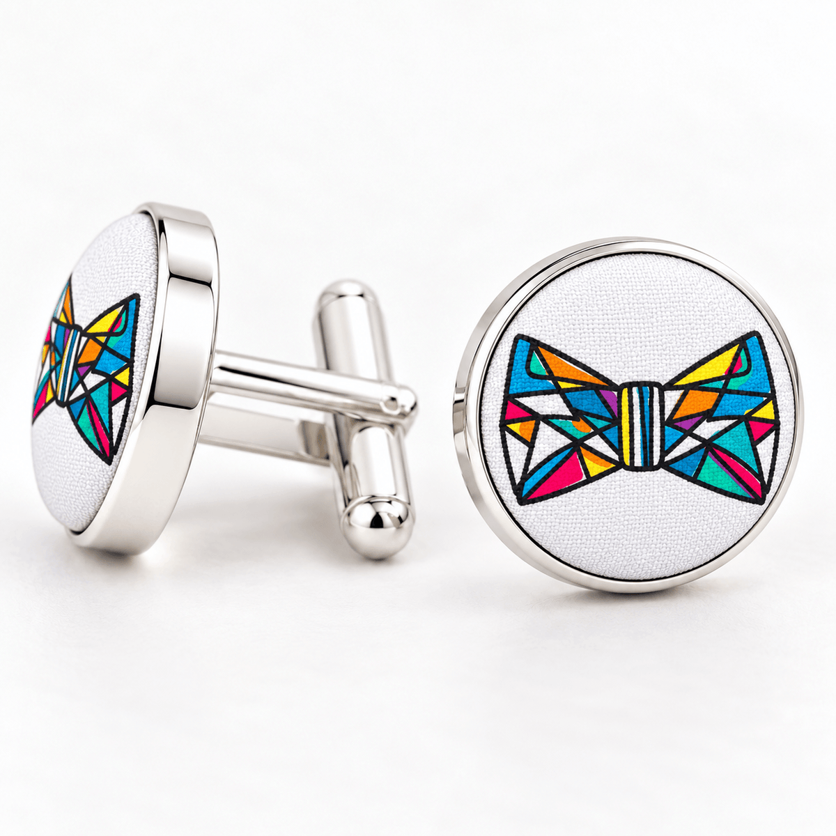A pair of round silver cufflinks on a white background. One cufflink is shown from the back with its silver bar and toggle, and the other is shown from the front featuring a colorful geometric bow tie design on a white textured background.