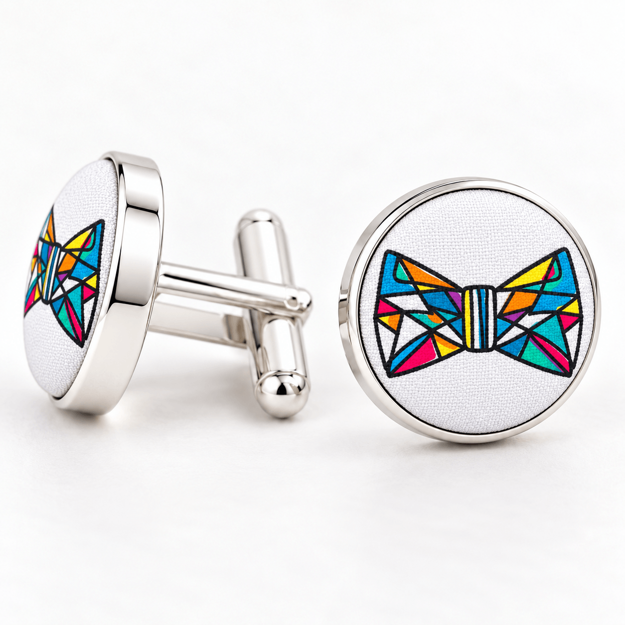 A pair of round silver cufflinks on a white background. One cufflink is shown from the back with its silver bar and toggle, and the other is shown from the front featuring a colorful geometric bow tie design on a white textured background.