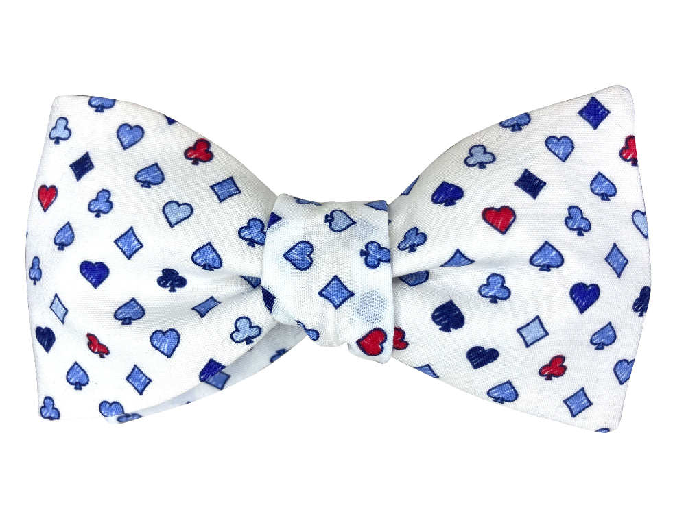 A white bow tie featuring a repeating pattern of blue and red playing card suits (hearts, spades, clubs, and diamonds)